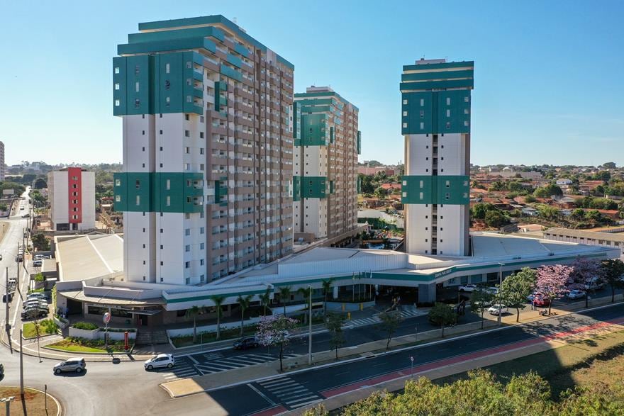A modern resort complex is presented with three tall buildings, featuring a mix of white and teal exteriors. The layout includes a large entrance area and surrounding greenery, with a view of the cityscape in the background, highlighting the vibrant neighborhood.