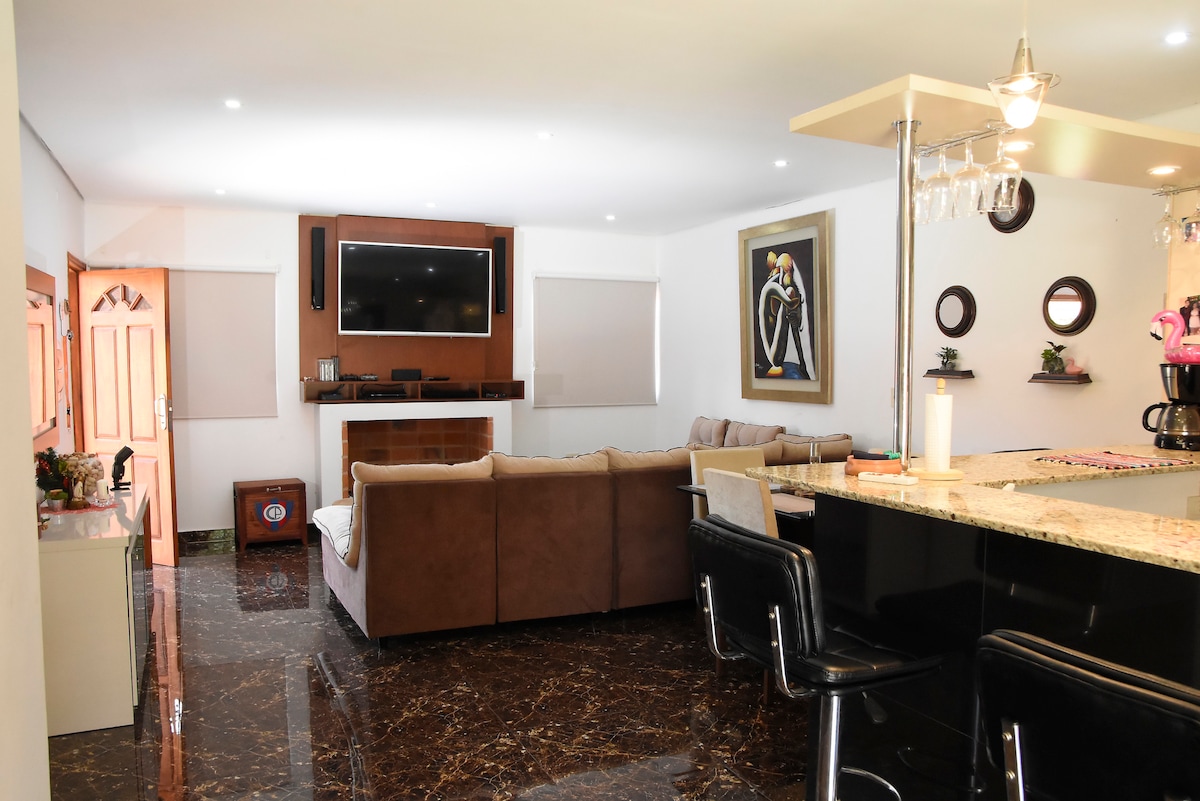 A spacious living area features a large, comfortable sectional sofa facing a modern television mounted above a decorative fireplace. The sleek kitchen bar with black seating contrasts with the elegant marble floor, while natural light fills the space through shaded windows.