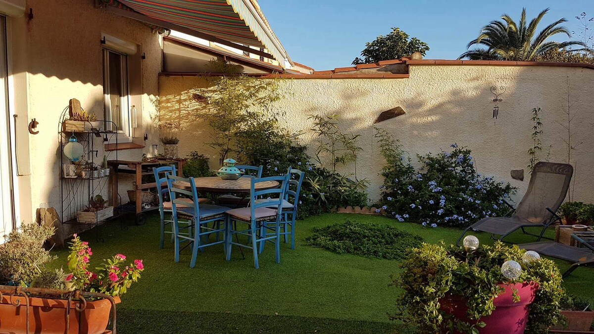 A green, enclosed garden features a seating area with a round table and six blue chairs. Lush plants, including flowers in pots, create a vibrant atmosphere. A lounge chair is positioned nearby, complementing the sunny ambiance. Sunlight casts soft shadows across the artificial grass.