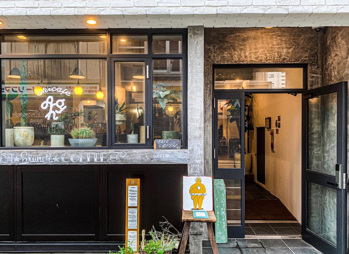 The entrance of akicafe inn is framed by large glass windows, allowing natural light to enter. The cozy café area features a welcoming decor, with playful artwork on the walls. A chalkboard sign indicatively displays the coffee offerings, inviting guests to explore.