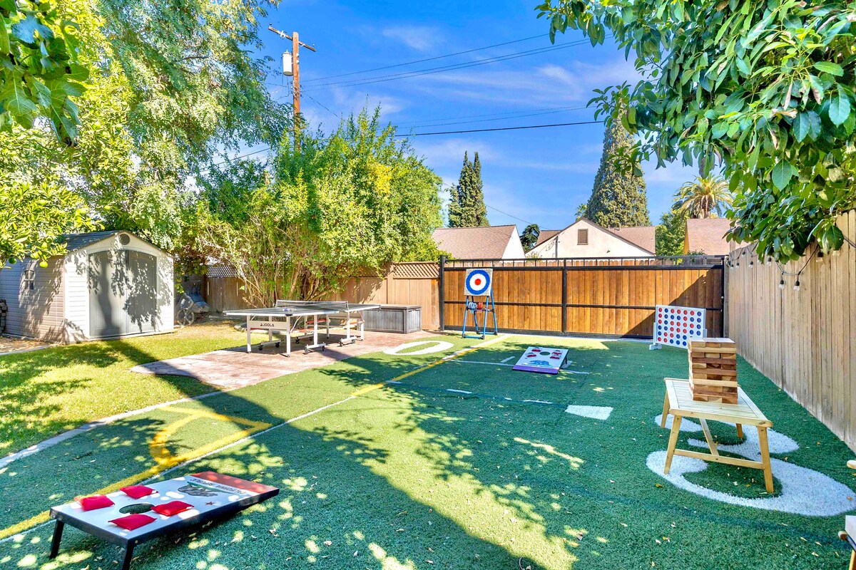 An expansive backyard features various outdoor games, including cornhole, ping pong, and life-size Jenga. Lush greenery surrounds the space, providing shade and a natural atmosphere. A charcoal grill is visible, promoting an inviting area for alfresco dining.