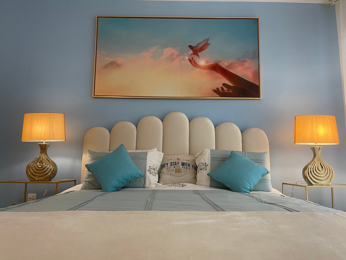 The sleeping area features a spacious bed with a soft headboard and decorative blue cushions. Two stylish lamps provide soft lighting beside the bed. A serene artwork depicting hands reaching for a dove is prominently displayed above the headboard, enhancing the room's calming ambiance.