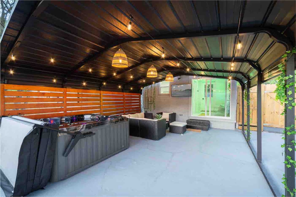 An outdoor covered patio features a large hot tub, comfortable seating arrangements, and ambient lighting from modern fixtures. The enclosed space includes wooden slats for privacy, while a mounted TV offers entertainment options. The area is designed for relaxation and social gatherings.
