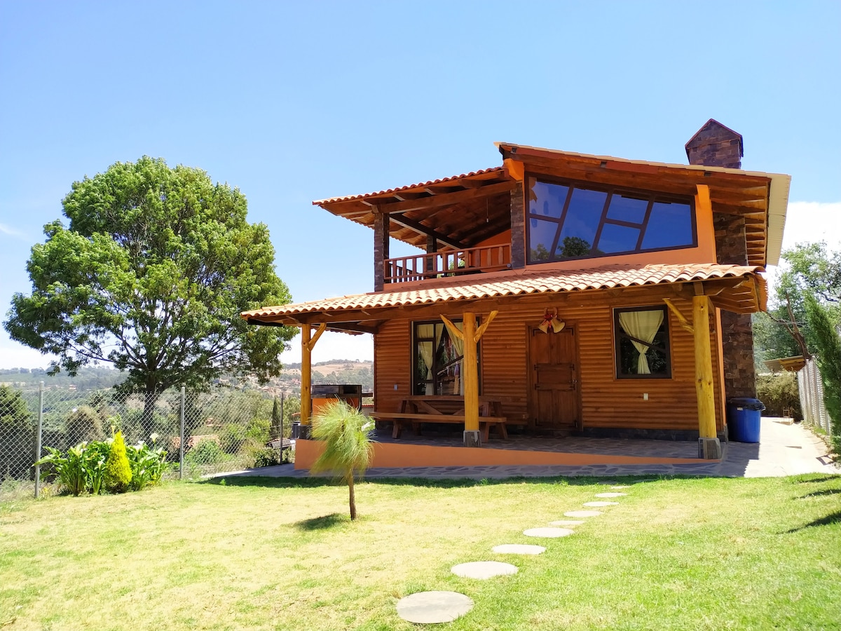 A spacious rustic cabin is presented, featuring large windows that allow natural light to fill the interior. A wooden porch with seating offers views of the surrounding greenery. Lush grass and a pathway of stepping stones lead to the cabin’s entrance.