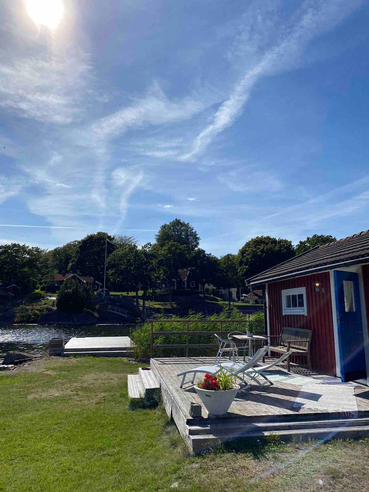 A red guesthouse is visible near a serene waterway, featuring a wooden deck with outdoor seating. Lush greenery surrounds the area, with a small dock accessible from the property. Bright blue skies fill the background, complemented by scattered clouds.