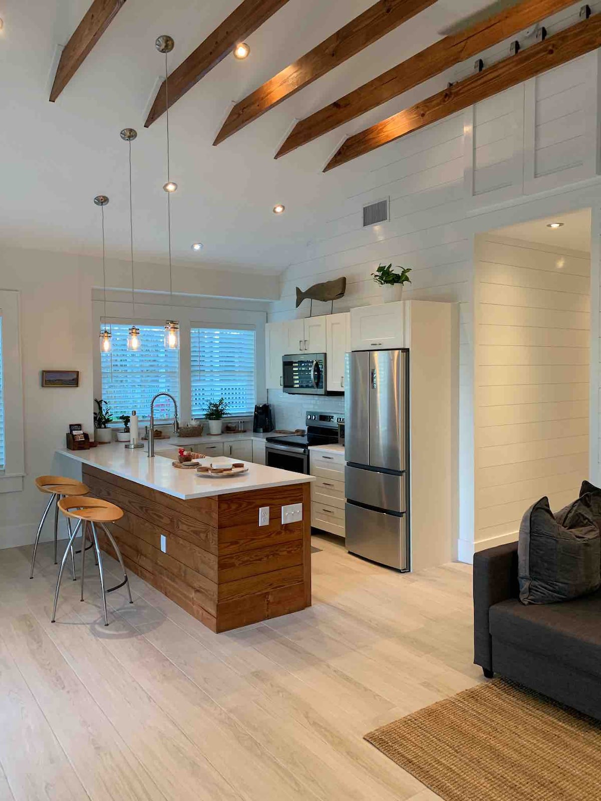 An open concept kitchen is highlighted by a large island with wooden accents and modern bar stools. Stainless steel appliances complement quartz countertops, while recessed lighting illuminates the space, which features white cabinetry and exposed wooden beams above.
