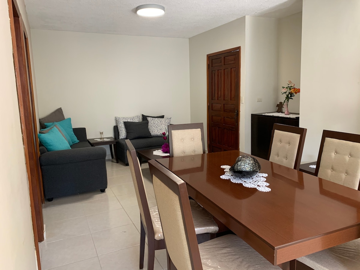 A spacious living and dining area is visible, featuring a well-maintained dining table with light-colored chairs. A comfortable sofa is present, adorned with teal cushions. Natural light brightens the space, complementing the neutral wall tones and wooden door.