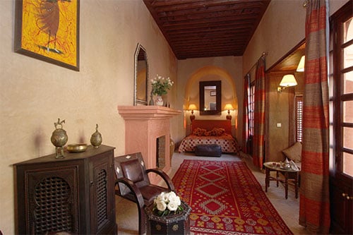 A welcoming suite features a cozy sitting area with two chairs and an ornate table. A spacious bed is situated in the background, adorned with patterned bedding. A decorative rug adds warmth to the stone floor, while a fireplace and large windows enhance the ambiance.