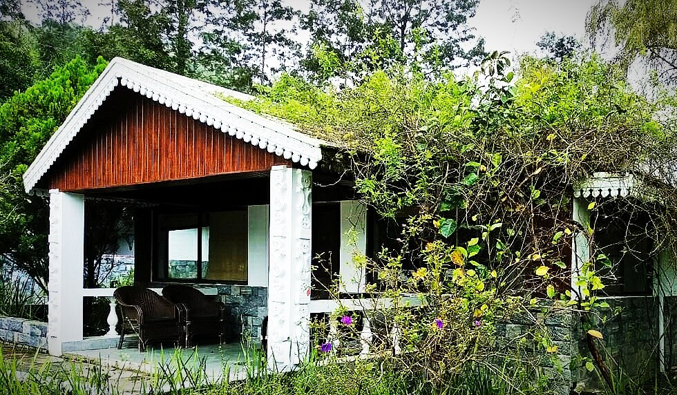 The cottage features a welcoming covered verandah, supported by decorative white columns, with a natural ambiance created by surrounding greenery. A cozy sitting area is visible, offering a connection to the tranquil outdoor environment.