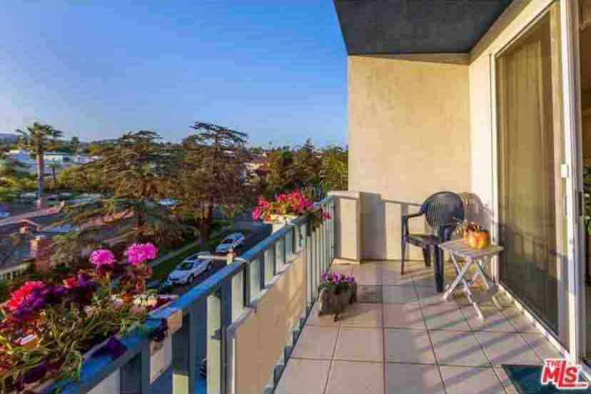 A private balcony features a small table and chair, surrounded by blooming flowers. The view includes distant trees and parked cars, with soft sunlight illuminating the space. The balcony's tiled floor enhances the outdoor experience.