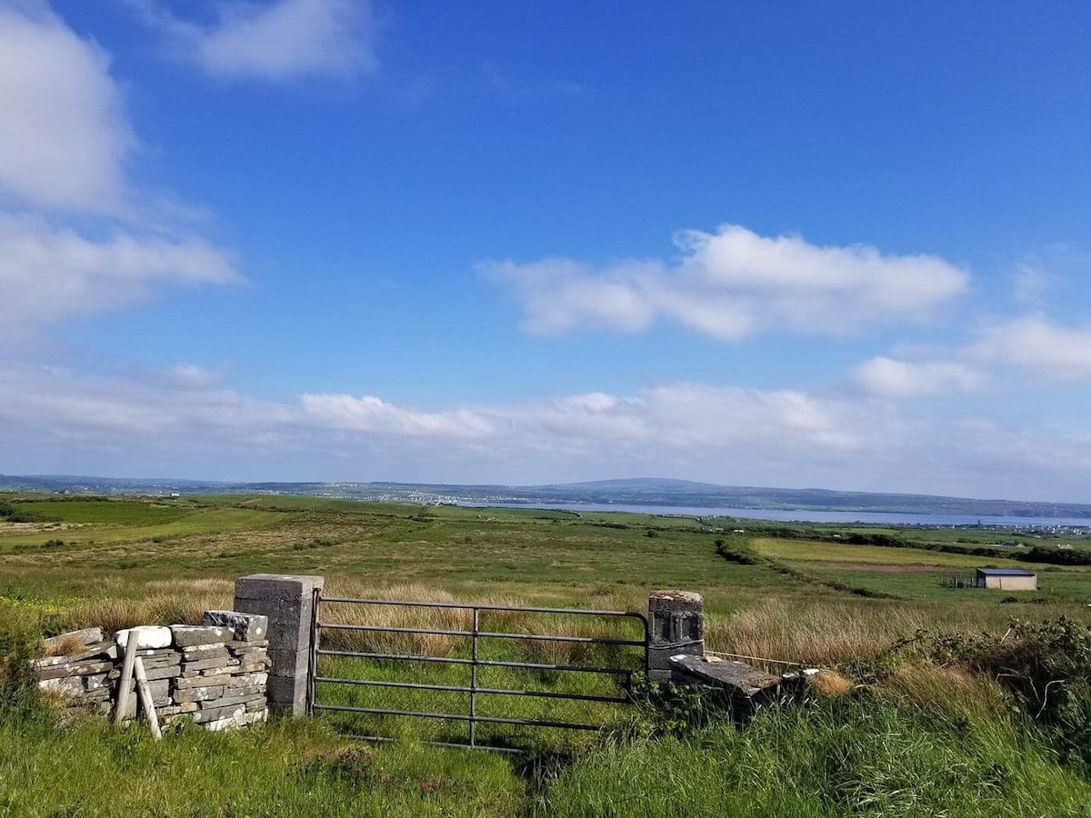 A wide-open landscape is captured, showcasing lush green fields under a bright blue sky. A rustic stone wall frames a metal gate leading towards a scenic view of the countryside and Liscannor Bay in the distance.