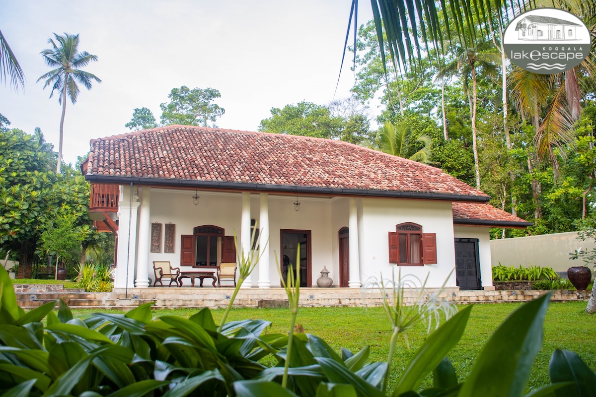 The colonial-style house features a sloped roof with red tiles and wooden shutters. Lush greenery surrounds the property, which includes a welcoming porch with seating. The symmetrical façade creates a balanced aesthetic against the backdrop of tropical trees.
