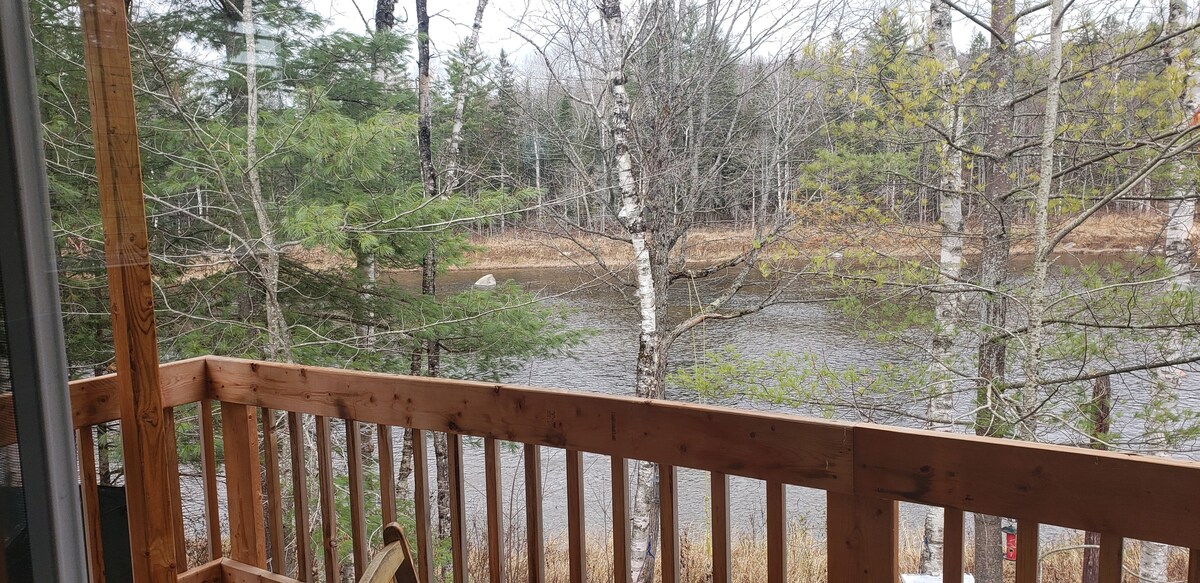 A wooden balcony offers a view of the calm river, surrounded by leafy trees and soft grasses. The natural scenery reflects tranquility, with the gentle flow of water visible in the background.