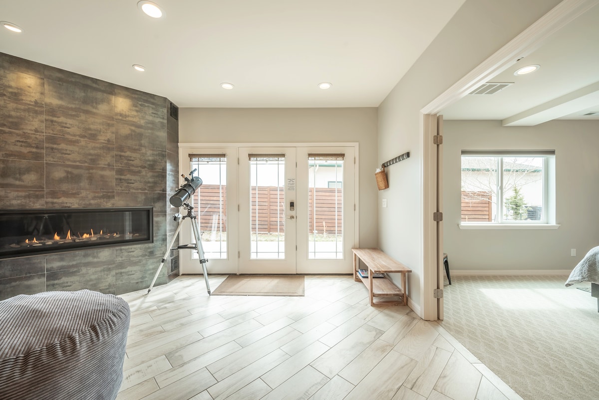 A spacious living area is highlighted by a modern gas fireplace along a textured wall. A telescope is positioned nearby, and natural light floods through glass doors that lead to a private outdoor space. A wooden bench adds to the comfort of the room.