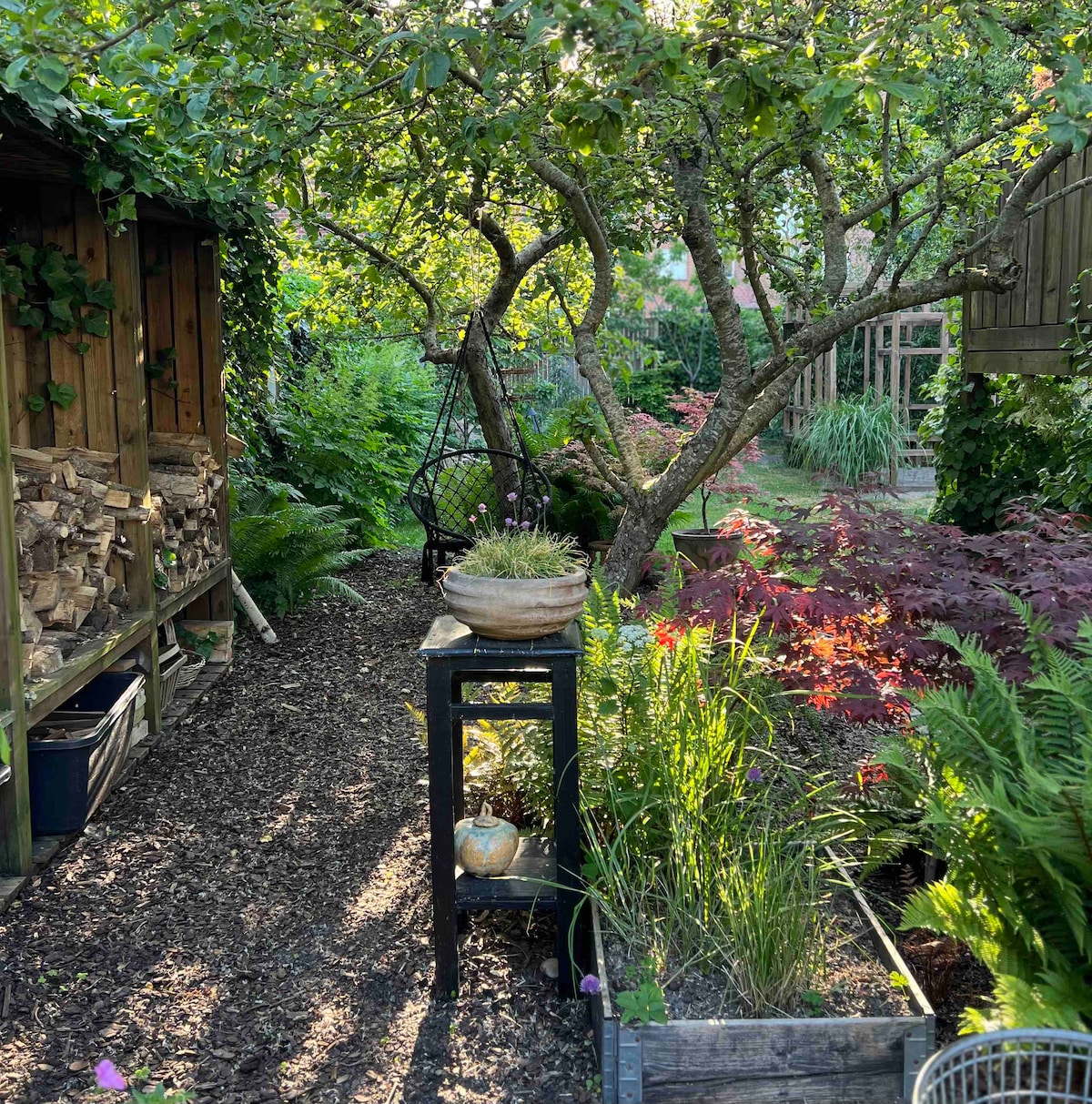 A serene garden path is framed by lush greenery and vibrant plants. A small table holds a decorative pot, while wooden storage for logs is positioned nearby. The area features dappled sunlight filtering through trees, creating a tranquil outdoor environment.