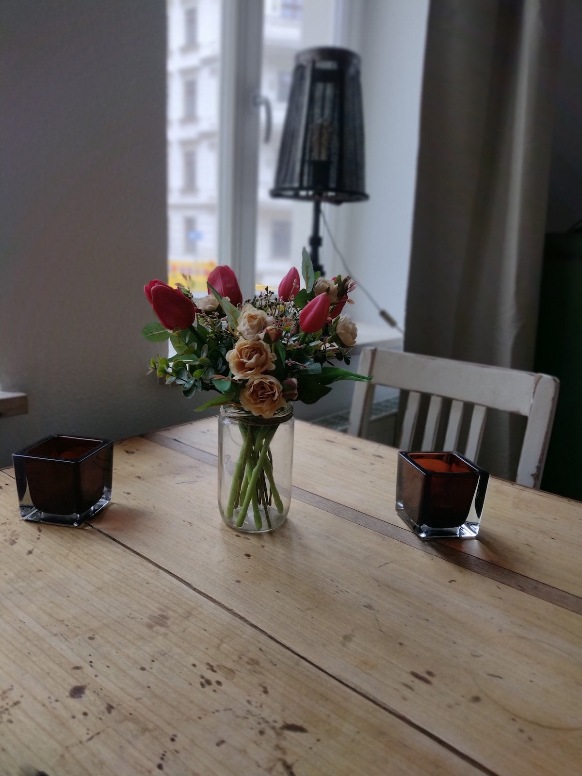 A wooden dining table is adorned with a vase holding an arrangement of artificial flowers, featuring roses and tulips. Two square candle holders are positioned on either side, adding decorative elements to the setting. Natural light filters through a nearby window.