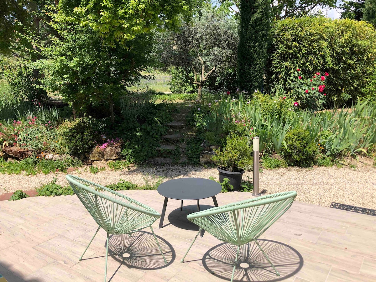 A tranquil outdoor terrace features two green chairs and a small black table, surrounded by a lush garden. Vibrant flowers and greenery create a serene environment, while a gravel path leads into the garden, inviting exploration.