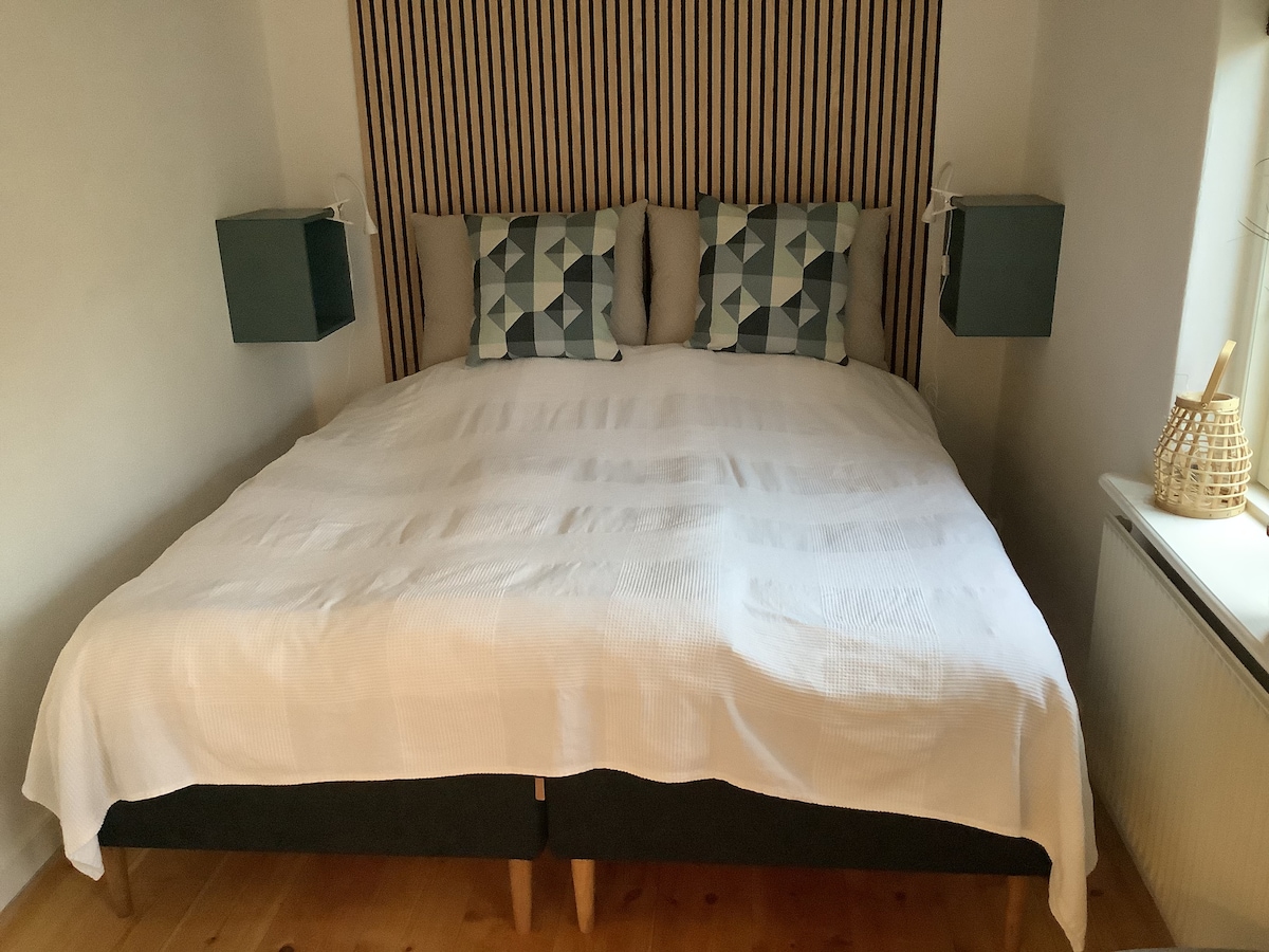 A spacious bed is positioned against a vertically slatted wooden headboard. Two bedside tables flank the bed, each with modern green lamps. The bed is made with a white coverlet, complemented by patterned pillows in shades of blue and grey.
