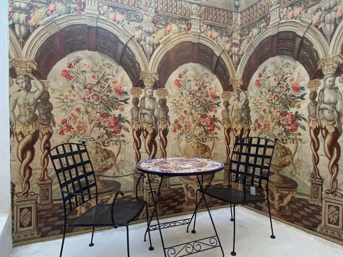 An outdoor patio space features a small round table decorated with a vibrant mosaic design, accompanied by two black metal chairs. The walls are adorned with an intricate mural depicting classical architecture and floral arrangements, creating an inviting atmosphere for relaxation.
