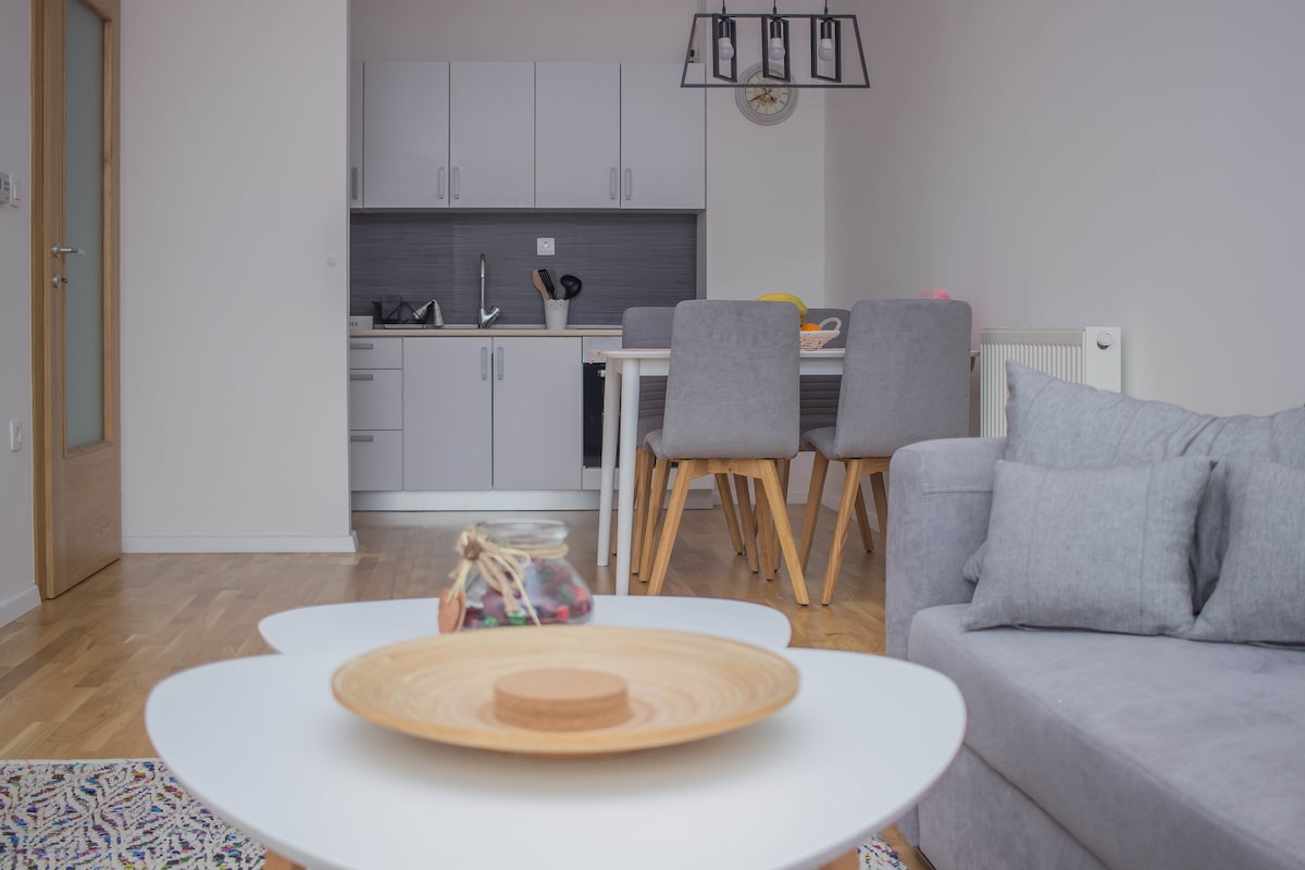 A modern living area showcases a light-colored, round coffee table in the foreground. A gray sofa sits comfortably beside it. The open kitchen features sleek cabinets and a dining table with gray chairs, creating a functional space for cooking and dining.
