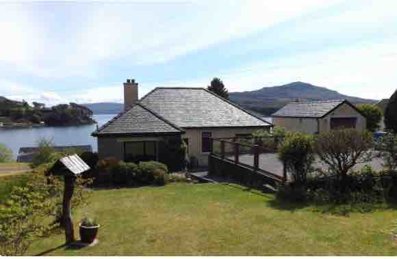 A single-story house is set amidst a well-maintained garden, featuring a mix of shrubs and grass. The backdrop includes the tranquil waters of Portree bay and rolling hills, providing a serene atmosphere. A patio area is visible, enhancing outdoor enjoyment.