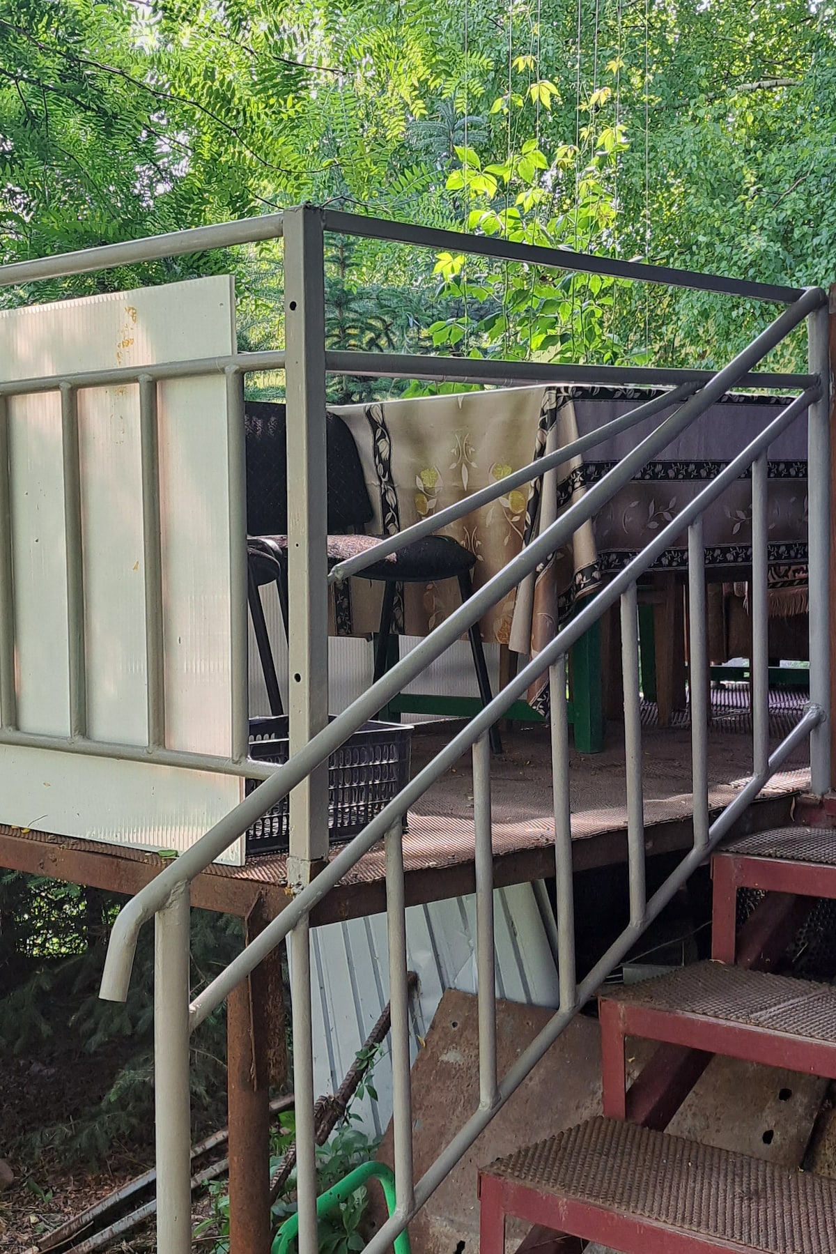 An outdoor balcony is shown, featuring a table with chairs surrounded by lush greenery. A set of stairs leads to the elevated platform, while a railing provides safety. Natural light filters through the trees, creating a serene setting.