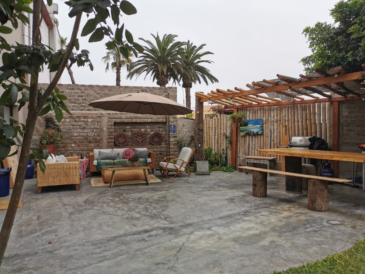 A spacious outdoor area features a shaded seating arrangement with a large umbrella, and a comfortable couch accompanied by a rocking chair. A wooden table made from logs offers a natural touch, while palm trees and greenery frame the space, creating a serene environment.