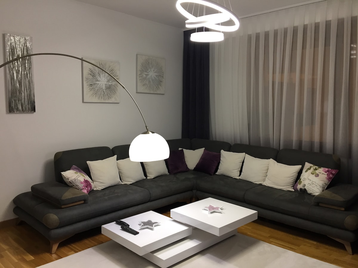 A spacious living area features a gray sectional sofa adorned with decorative pillows in soft colors. A modern coffee table with a unique design sits in the center, and a contemporary light fixture hangs above, complemented by sheer curtains that allow natural light to filter in.