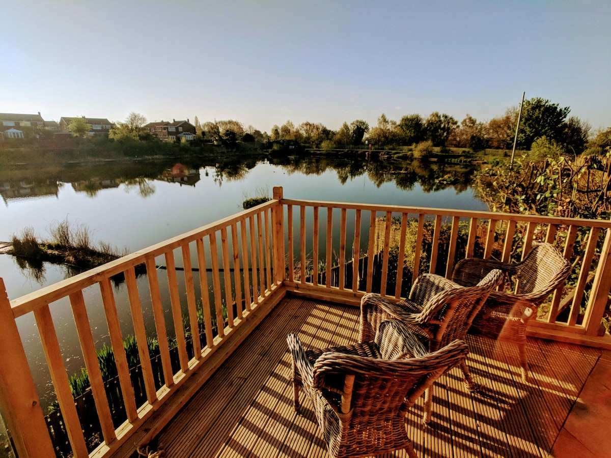A wooden deck overlooks a serene lake, with two wicker chairs positioned invitingly. The surrounding landscape features lush greenery and reflections of the shoreline, creating a peaceful atmosphere for outdoor relaxation.