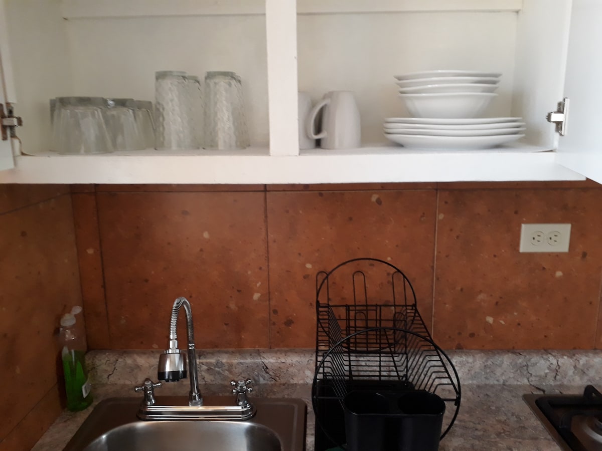 The kitchen area features a stainless-steel sink positioned beneath a cabinet. Clear glassware and white dinnerware are neatly organized on the shelves above. A dish drying rack is visible next to the sink, with a small container for utensils. The countertop is made of a speckled design.