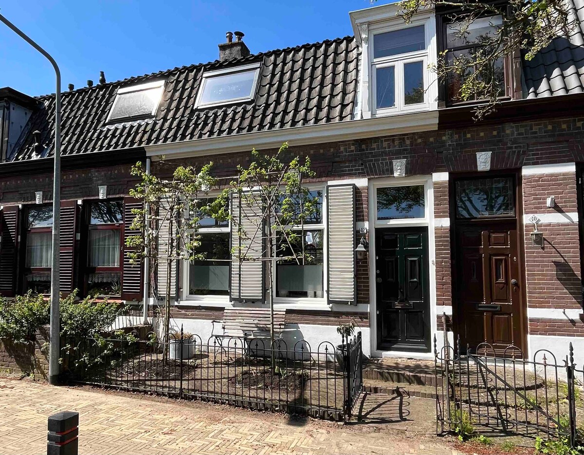 A charming row house is presented, featuring a traditional brick facade with decorative elements. Windows are adorned with shutters, and a small garden area is visible in front, enclosed by a low fence. The entrance door is framed by a symmetrical arrangement of plants.