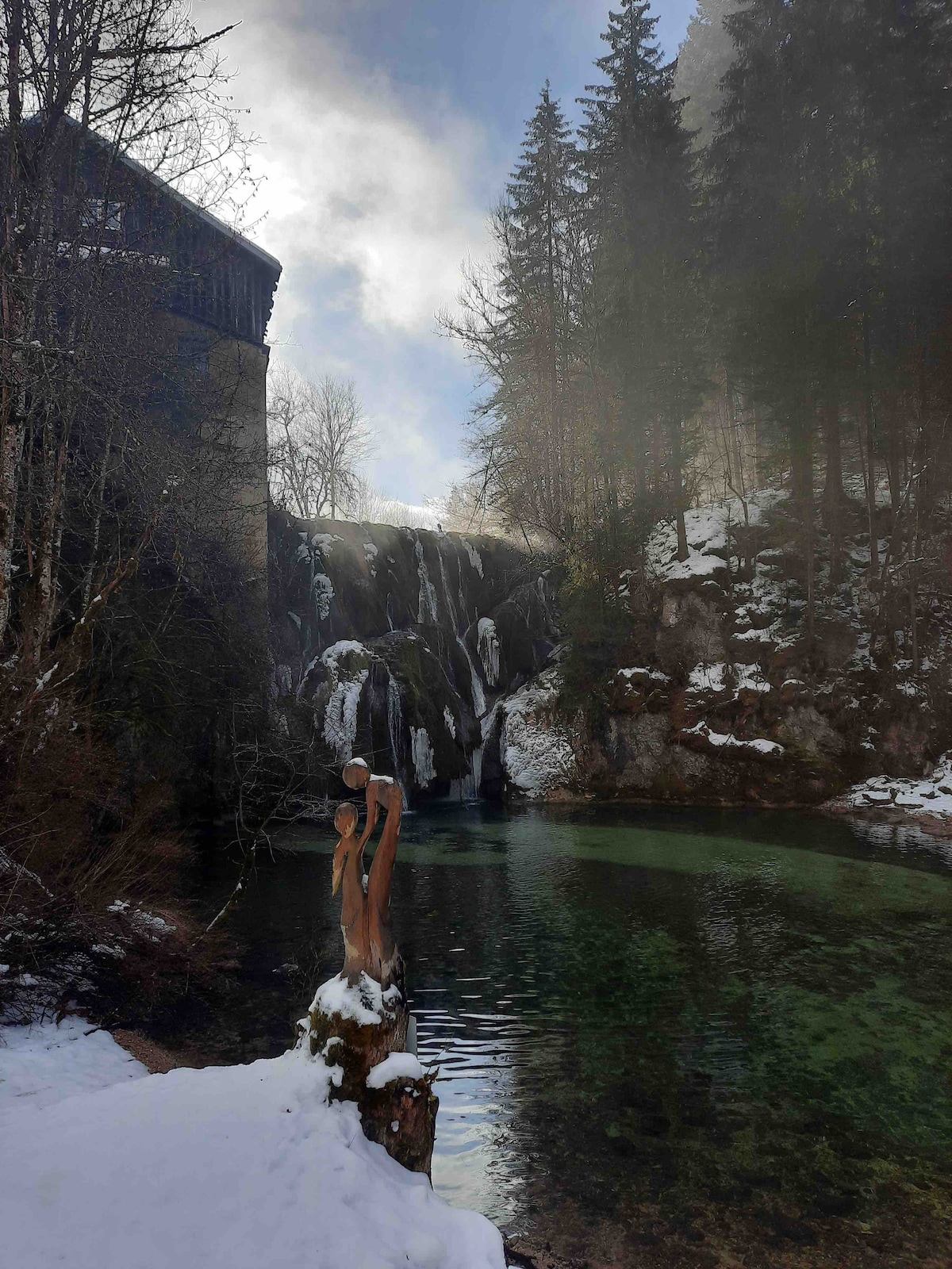 A waterfall cascades into a serene pool, surrounded by a snowy landscape and evergreen trees. The water appears clear and reflects the sky, while mist rises gently above the falls, creating a peaceful atmosphere. A partially visible wooden structure is situated in the background.