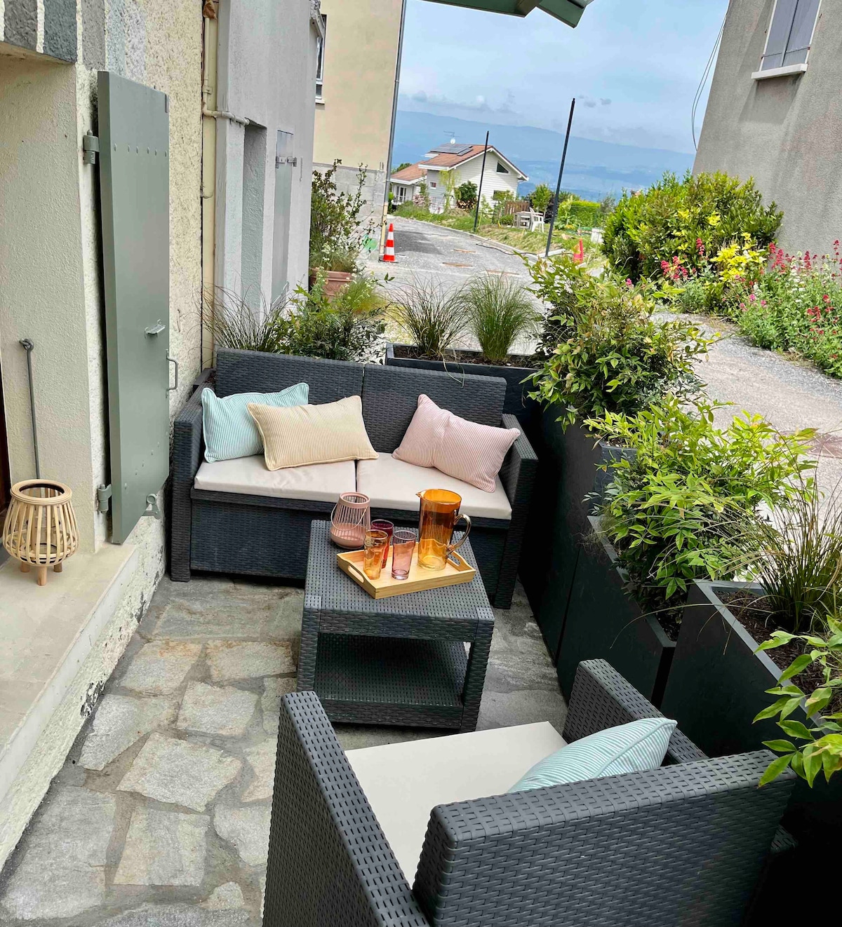 A small terrace area features a seating arrangement with soft cushions in muted tones. A coffee table is adorned with decorative glassware. Lush greenery and potted plants surround the space, enhancing the outdoor ambiance while providing privacy.