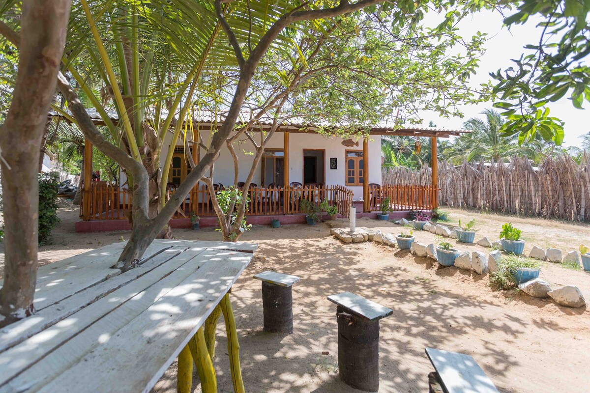 A shaded garden area features a rustic wooden table surrounded by two cylindrical stools. The nearby villa, with a porch and large windows, is partially visible through lush greenery, providing a serene outdoor space shaded by trees.