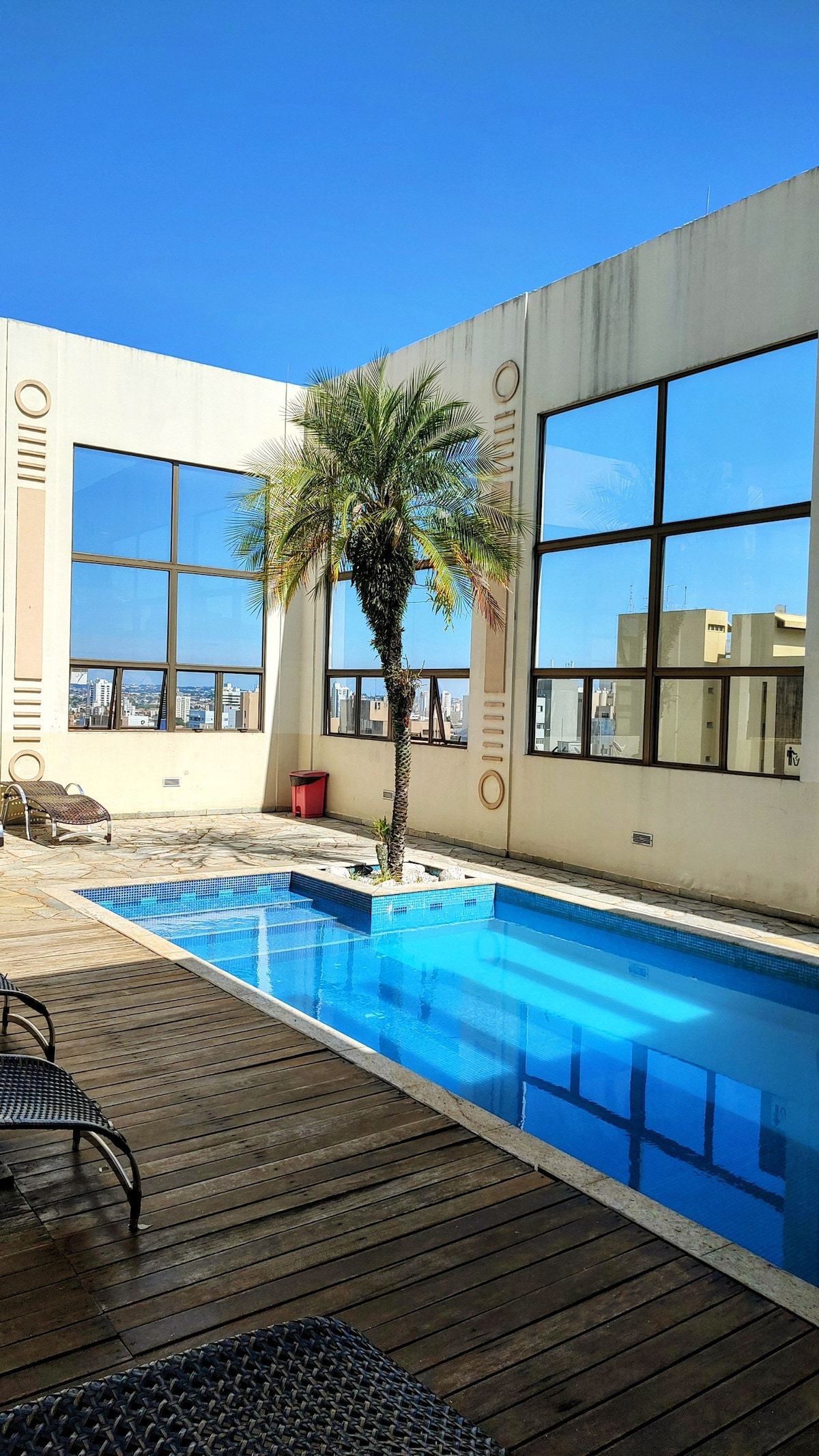 A rooftop pool area is presented, featuring a rectangular pool surrounded by wooden decking. Large windows allow natural light to flood the space. A palm tree is positioned next to the pool, providing a touch of greenery to the setting. Lounge chairs are arranged nearby.