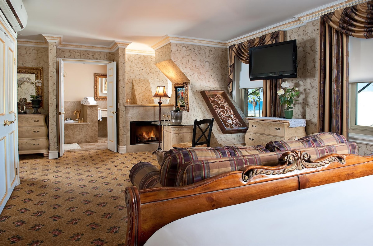 A spacious room features elegant Victorian design with a wooden bed, decorative furniture, and a flat-screen TV. A cozy fireplace sits to one side, while windows provide natural light. An open doorway reveals a well-appointed bathroom with plush towels visible.