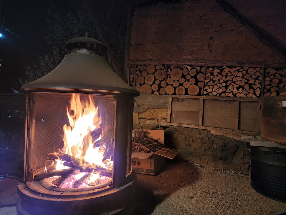 A fireplace is prominently featured, with flames dancing inside a metal frame. Stacks of firewood are neatly arranged against the back wall, providing an inviting ambiance. The surrounding area showcases a rustic interior, highlighted by natural materials and warm, subtle lighting.