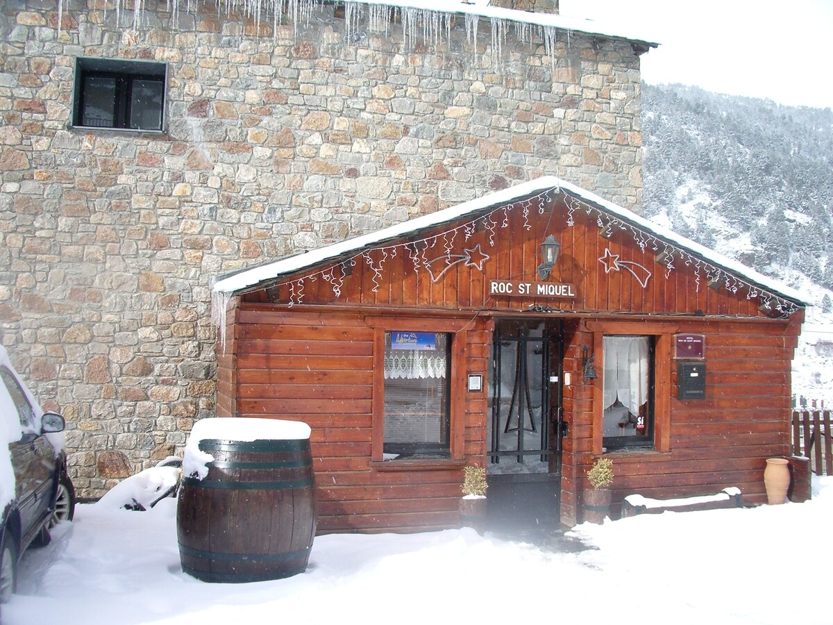 The exterior of the Hotel Roc de St Miquel is featured, showcasing a stone and timber facade accented by decorative elements. A snow-covered landscape surrounds the building. An entrance is visible, flanked by a large wooden barrel. Icicles hang from the roof edges.