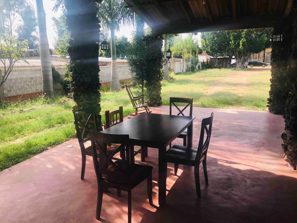 An outdoor patio is shown, featuring a wooden dining table surrounded by six wooden chairs. Natural light filters through the trees, creating a serene atmosphere. The expansive green yard is visible in the background, providing a spacious and open environment.