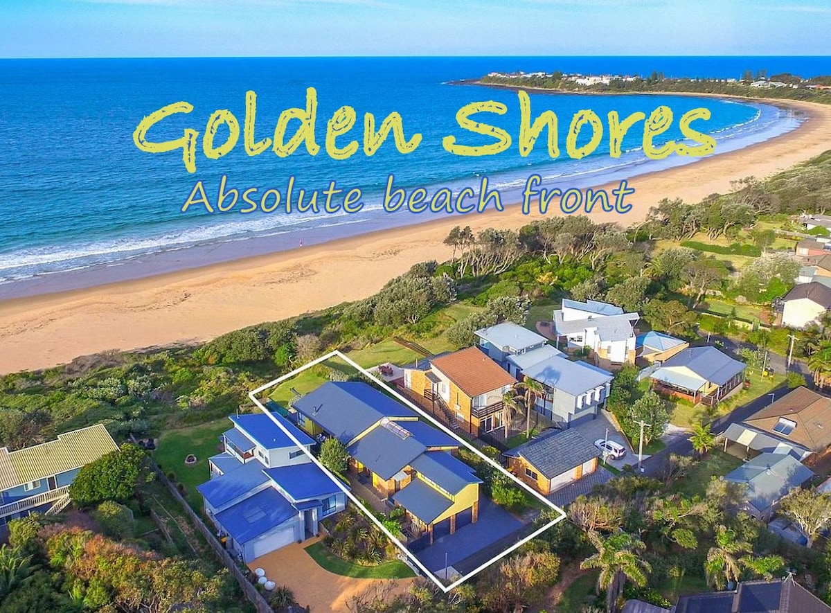 An aerial view showcases the beach front location of the house, surrounded by lush greenery and neighboring residences. The golden sand beach and sparkling blue ocean stretch out prominently in the background, highlighting the proximity to the shore.
