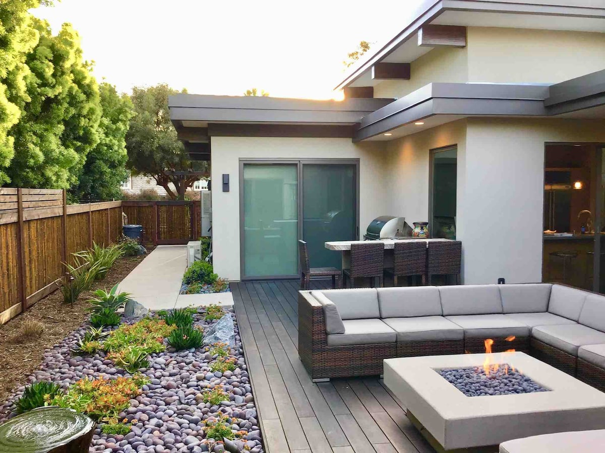 An outdoor space features a modern seating arrangement surrounding a gas fire pit. A well-maintained garden with colorful plants is visible, along with a dining area equipped with a grill. The natural stone pathway leads towards a gated area showcasing trees and greenery.