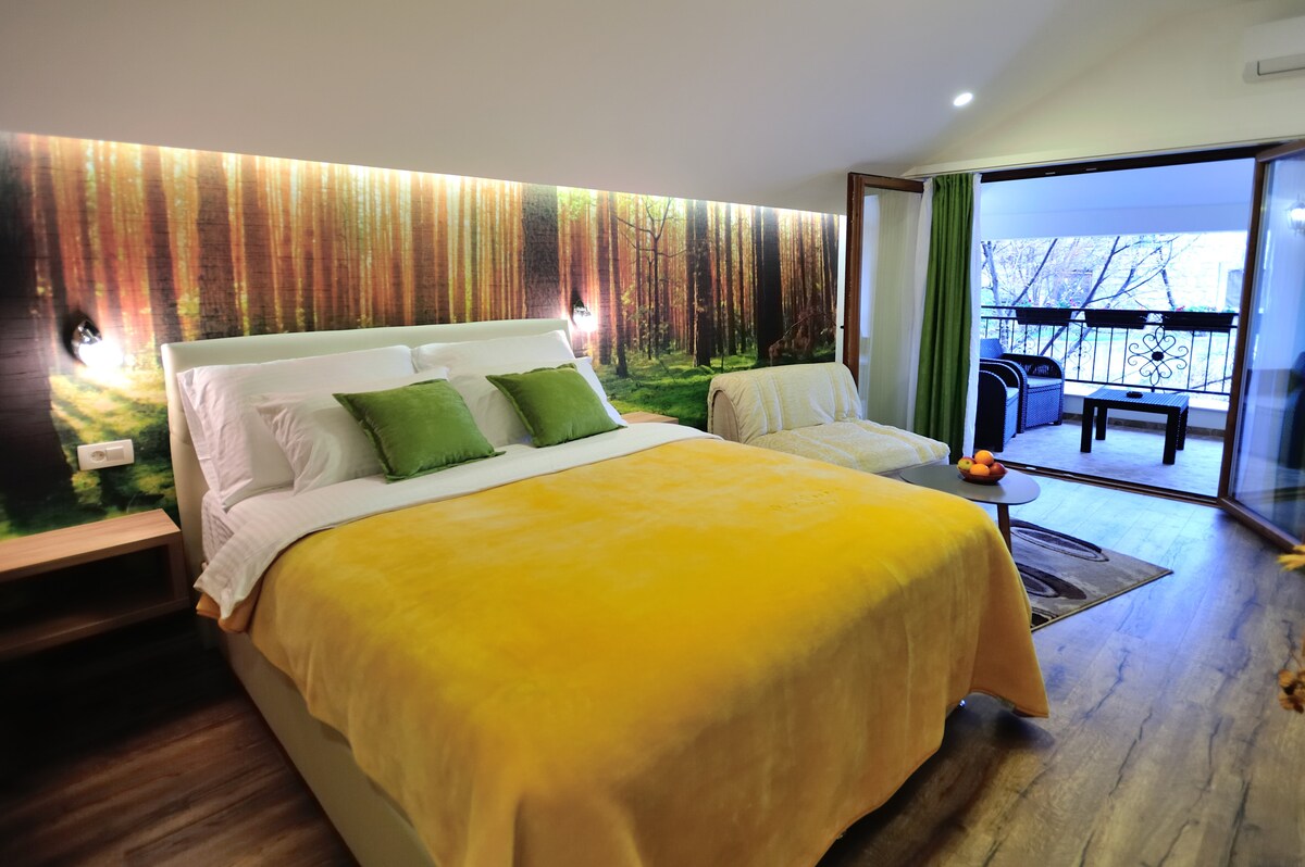 A cozy room features a spacious bed topped with soft pillows, complemented by a bright yellow blanket. A calming forest mural creates a tranquil ambiance. A small table holds fresh fruit, and a balcony with seating is accessible through glass doors.