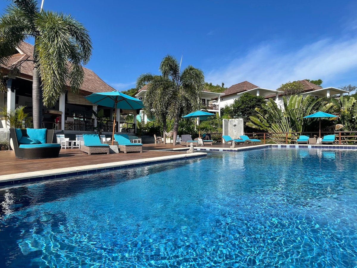 An expansive infinity pool reflects the clear blue sky, surrounded by comfortable lounge chairs and vibrant umbrellas. Lush tropical plants create a serene backdrop, while a modern restaurant structure is visible in the distance, providing a tranquil setting for relaxation.