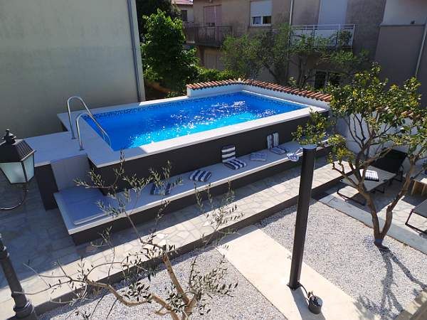 A swimming pool is situated in a landscaped area, surrounded by serene greenery. Lounge chairs are placed along one side of the pool. The water's surface reflects sunlight, and the pool is designed with steps for easy access.