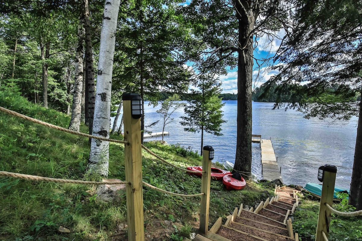 A wooden staircase is lined with rope, leading down through lush greenery to a serene lakefront. Kayaks in vibrant colors are positioned at the water's edge, while trees provide shade and depth to the natural setting.