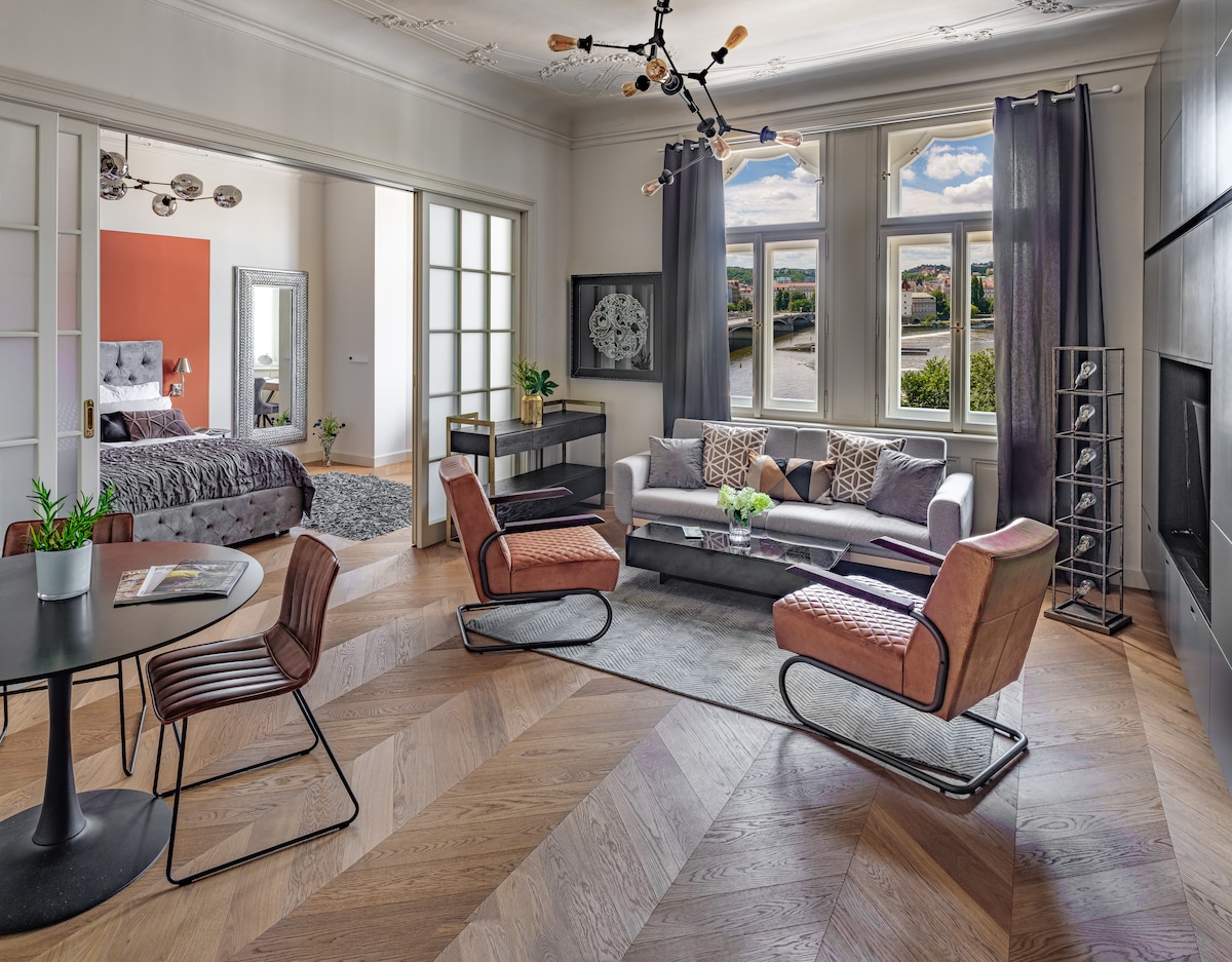 A spacious living area is illuminated by large windows, offering a view of the river. The room features a comfortable seating arrangement with two stylish chairs and a modern sofa. A round dining table is positioned nearby, complemented by elegant decor elements.