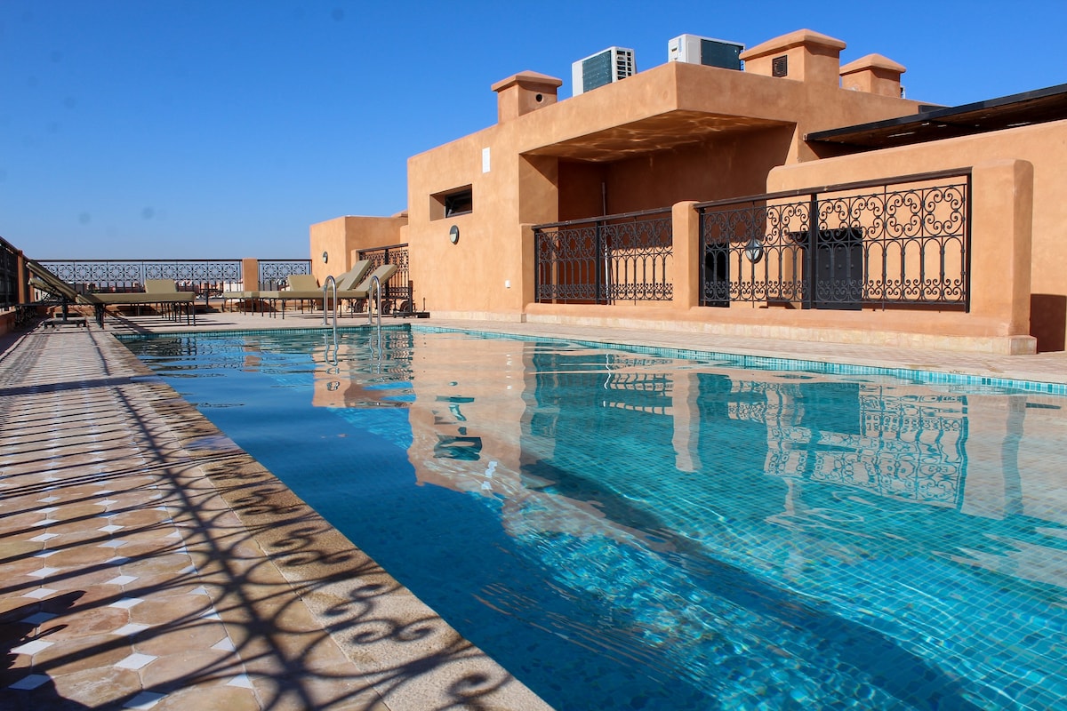 The image illustrates a tranquil swimming pool area complemented by a decorative tile border. Sun loungers are arranged nearby, with clear blue skies overhead. The architectural design features reddish-brown walls and intricate wrought-iron detailing, enhancing the serene outdoor space.