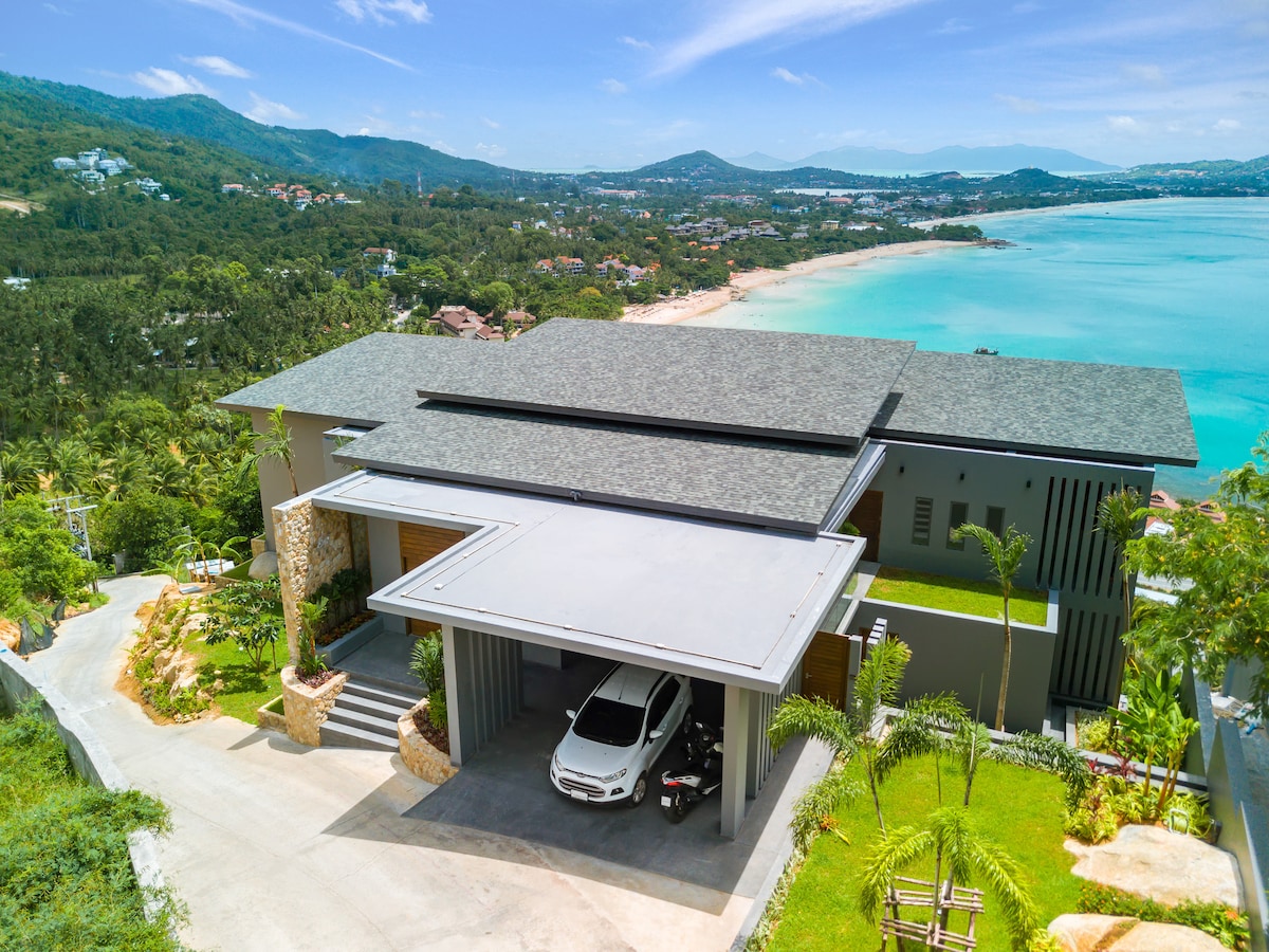 A modern villa is shown from an elevated angle, featuring a spacious carport beneath a stylish roof. Lush greenery surrounds the property, while the sparkling coastline of Chaweng Noi is visible in the background, revealing the ocean's blue hues and the distant hills.