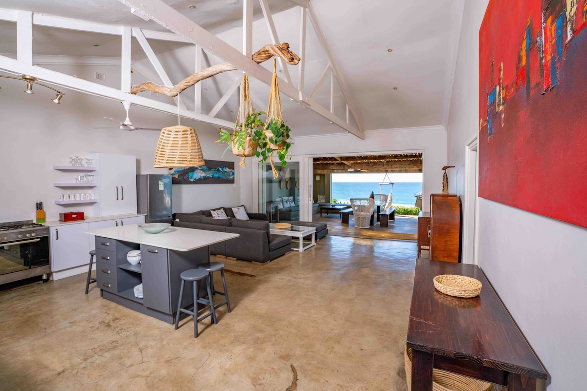 An open-plan living area features a modern kitchen with sleek cabinetry and a spacious island. A relaxed seating arrangement faces large windows, offering ocean views. Natural elements, such as hanging light fixtures and a wall adorned with artwork, enhance the coastal aesthetic.
