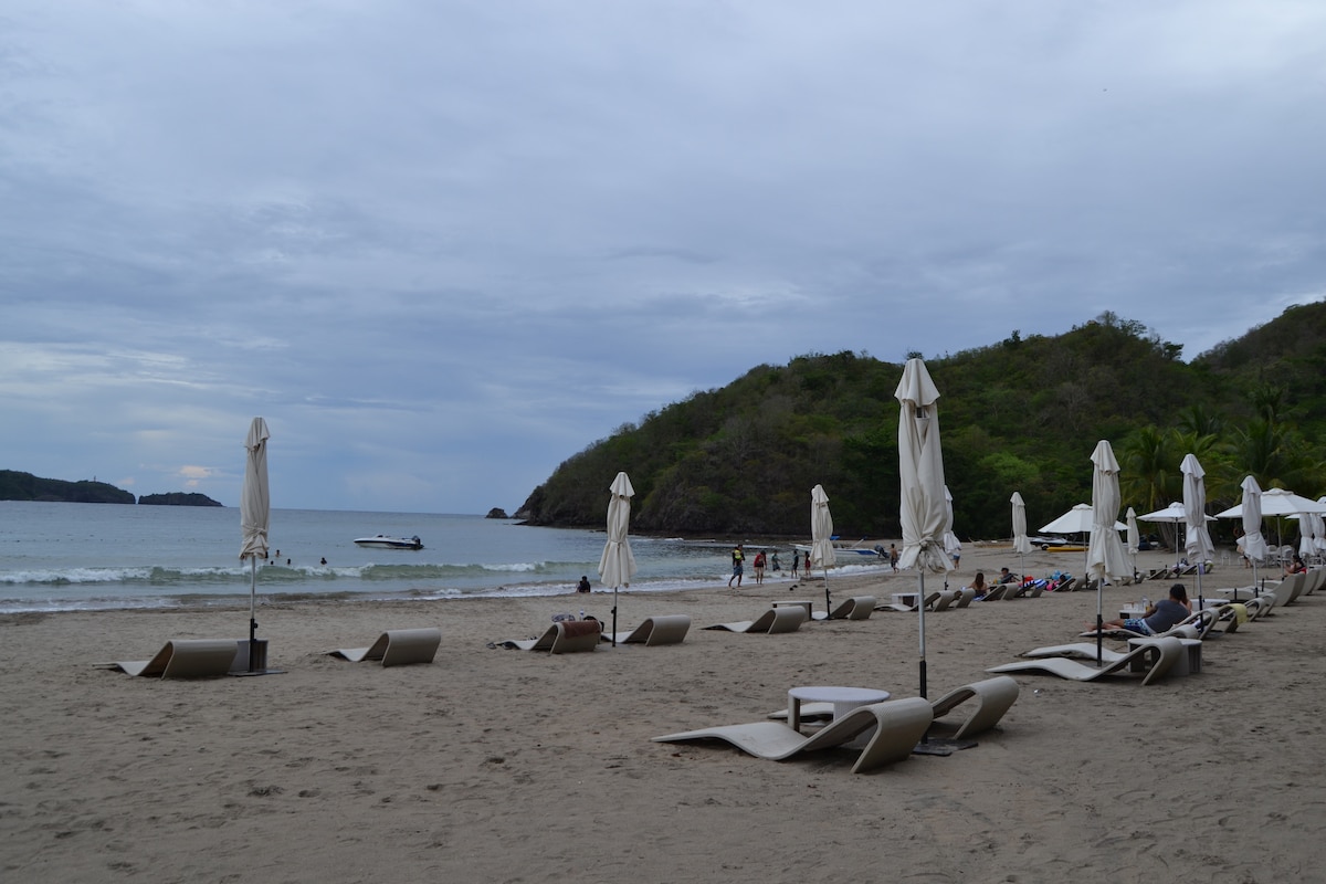 A sandy beach is lined with sun loungers and umbrellas, creating a relaxed setting. Gentle waves are observed lapping at the shore, with people enjoying the water in the distance. Lush greenery outlines the beach, enhancing the natural beauty of the area.